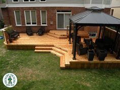 a wooden deck with chairs and a gazebo in the middle of grass next to a brick building