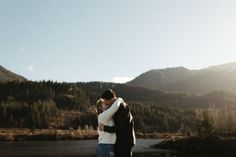 two people embracing each other in front of a lake and mountains with the sun shining on them