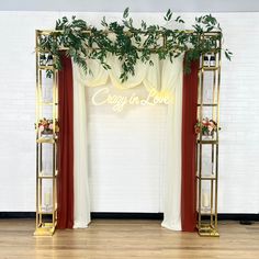 a white and gold wedding arch decorated with greenery, red drapes, and neon sign that says crazy in love