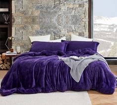 a bed with purple comforter and pillows in front of a stone wall