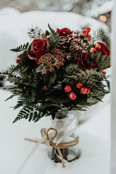 a bouquet of red flowers in a glass vase on the snow covered ground with ribbon tied around it