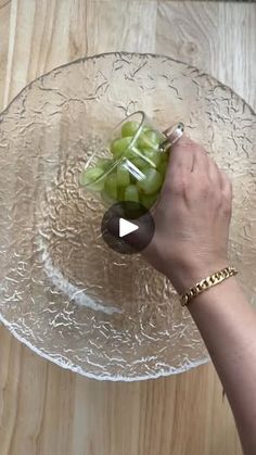 a glass plate with grapes on it being held by a person's hand over a wooden table