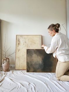 a woman kneeling down next to a painting on a bed with white sheets and pillows