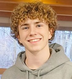 a young man with curly hair wearing a gray hoodie and smiling at the camera