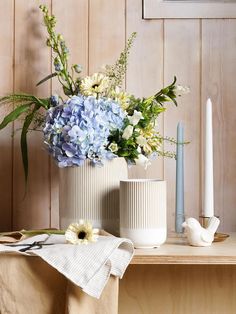 two white vases with blue flowers and greenery sit on a table next to a candle