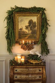 a painting hanging on the wall above a dresser with candles and greenery around it