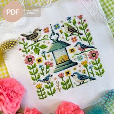 a cross stitch pattern with flowers and birds on it, next to a small bird feeder