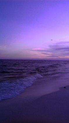 a purple sky over the ocean with waves coming in to shore and an airplane flying overhead