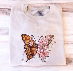 a white t - shirt with an orange butterfly and pink flowers on the front, sitting on a wooden surface