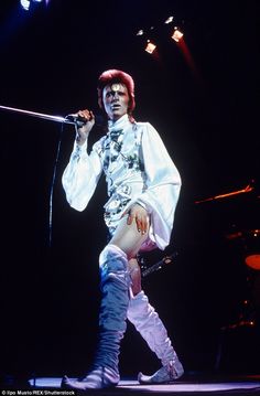 the man is dressed in all white and holding a microphone while standing on stage with his legs crossed