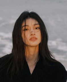 a woman with long black hair standing in front of the ocean and looking off into the distance