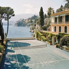 a tennis court in front of an ocean side house with trees and bushes around it