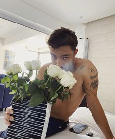 a shirtless man holding a vase with white roses in it and looking at the camera