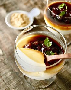 two glasses filled with dessert sitting on top of a wooden table next to spoons