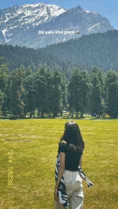 a woman standing on top of a lush green field next to a forest covered mountain