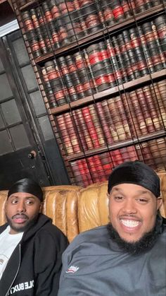 two men sitting on top of a couch in front of bookshelves filled with old books