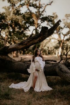 a pregnant woman in a white dress poses for a photo by a fallen tree trunk