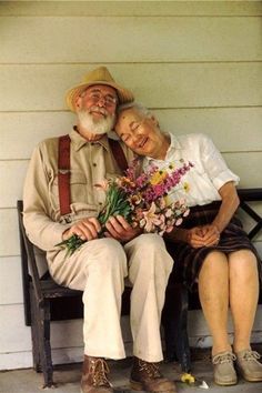 an old man and woman sitting on a bench next to each other with flowers in their hands