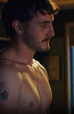 a shirtless man with a cross tattoo on his left arm and chest, looking off into the distance