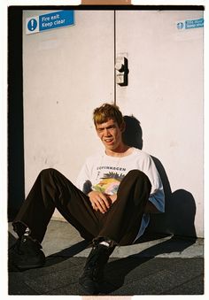 a young man sitting on the ground with his legs crossed and wearing black shoes, smiling at the camera