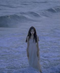 a woman standing in the ocean with her head turned to the side, wearing a long white dress