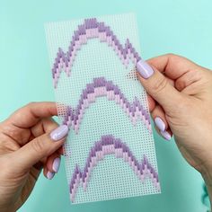 two hands holding up a piece of cross - stitch paper with pink and purple designs on it