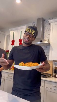 two men standing in a kitchen holding plates with food on them and one has his hands out to the camera