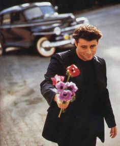 a man walking down the street with flowers in his hand and an old car behind him