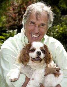 an older man holding a small dog in his arms and smiling at the camera with trees behind him