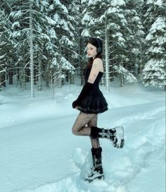 a woman in black dress and boots standing on snow covered ground with trees behind her