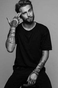 a man with tattoos sitting on a stool holding his hand up to his face and making the peace sign