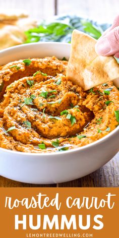 a hand dipping hummus into a white bowl with tortilla chips on the side