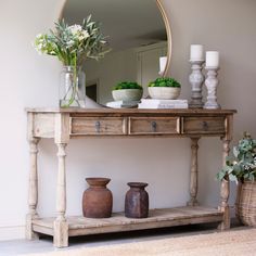 a wooden table with vases and flowers on it next to a mirror, candle holders and potted plants