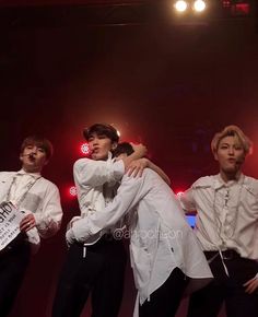 four men in white shirts and black pants hugging each other on stage with red lights behind them