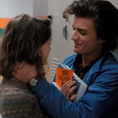 a man and woman standing next to each other in front of a door holding an orange book