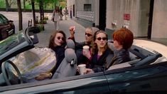 four women sitting in the back seat of a convertible car drinking coffee and talking to each other