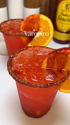 two glasses filled with red liquid and orange slices on top of each other next to bottles