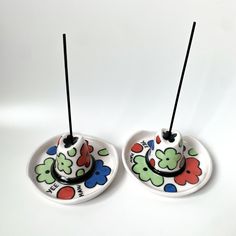 two small dishes with black sticks sticking out of them on a white surface, one is decorated with colorful flowers and the other has an upside down design