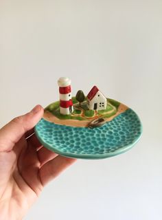 a hand holding a small plate with a house and lighthouse on it in front of a white background