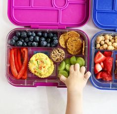 a child's hand reaching for an open lunch box filled with fruits and vegetables