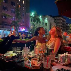 two women taking pictures with their cell phones at an outdoor dining table in the city