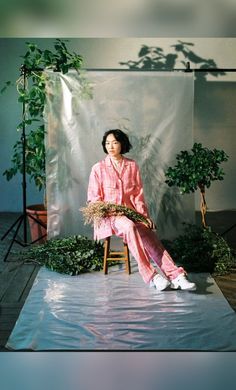 a woman sitting on top of a chair in front of a white backdrop and potted plants