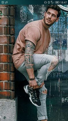 a man leaning against a brick wall with tattoos on his arm and leg, wearing sneakers