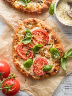 two pizzas with tomatoes, basil and cheese on paper next to some dipping sauce