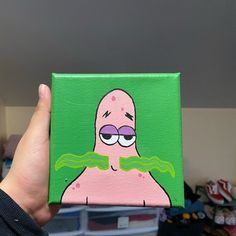 a hand holding up a small square painting of a pink cartoon character with green eyes