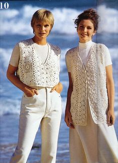 two women standing next to each other in front of the ocean wearing sweaters and pants