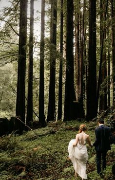 a bride and groom walking through the woods