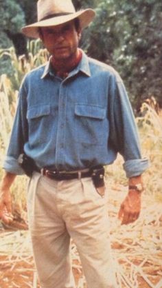 an older man standing in a field wearing a hat and blue shirt with his hands on his hips