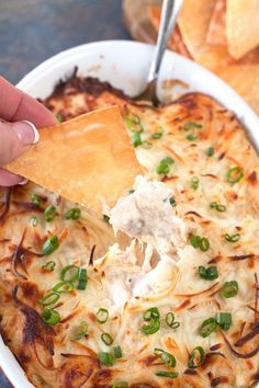 a hand holding a tortilla chip over a casserole dish with cheese and green onions