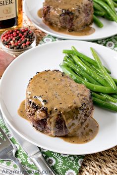 two white plates topped with meat covered in gravy next to green beans and a bottle of wine